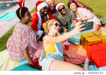 Happy diverse friends with santa hats having picnic and taking selfie in garden at christmas Happy diverse friends with santa hats having picnic and taking selfie in garden at christmas 95649135