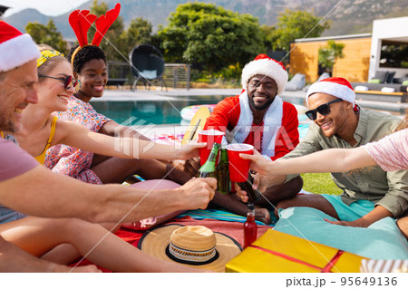 Happy diverse friends with santa hats having picnic in garden at christmas Happy diverse friends with santa hats having picnic in garden at christmas 95649136
