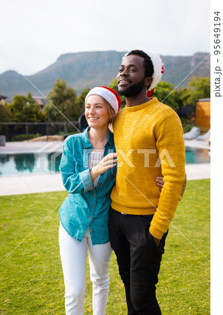 Vertical image of happy diverse couple celebrating christmas standing outdoors in santa hats 95649194