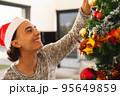 Happy biracial man wearing santa claus hat, decorating christmas tree in living room 95649859