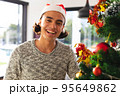 Happy biracial man wearing santa claus hat, decorating christmas tree in living room 95649862