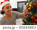 Happy biracial man wearing santa claus hat, decorating christmas tree in living room 95649863