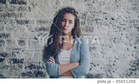 Portrait of angry young lady looking at camera, frowning and expressing disappointment, anger and disapproval standing with arms crossed with nice brick wall in background. Portrait of angry young lady looking at camera, frowning and expressing disappointment, anger and disapproval standing with arms crossed with nice brick wall in background. 95651208