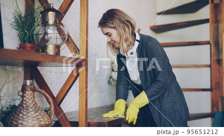 Professional housekeeper is dusting furniture with wet cloth, woman is wearing protective gloves, casual clothing and headphones. Housework, interiors and people concept. Professional housekeeper is dusting furniture with wet cloth, woman is wearing protective gloves, casual clothing and headphones. Housework, interiors and people concept. 95651212