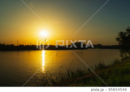 Sunset on the background of the river. The orange sun sets over the high-voltage power lines. In the foreground a bank of green grass. Sunset over the river in a small town. Sunset on the background of the river. The orange sun sets over the high-voltage power lines. In the foreground a bank of green grass. Sunset over the river in a small town. 95654549