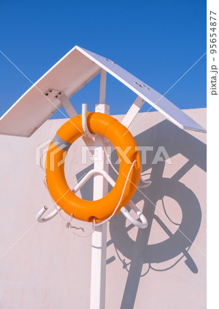Orange lifebuoy on beach by sea. Safety equipment for rescuing people on seashore. Orange lifebuoy on the pier of blue sky and azure sea Orange lifebuoy on beach by sea. Safety equipment for rescuing people on seashore. Orange lifebuoy on the pier of blue sky and azure sea 95654877