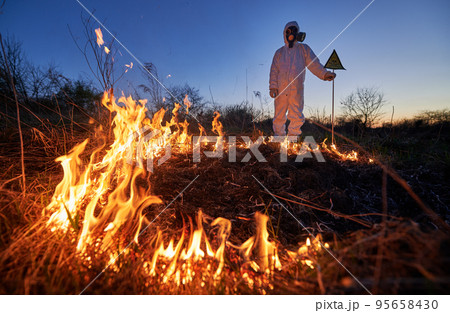 Firefighter ecologist working in field with wildfire at night. Man in suit and gas mask near burning grass with smoke, holding warning sign with skull and crossbones. Natural disaster concept. 95658430