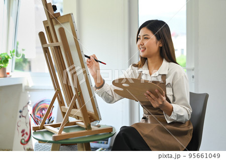 Pleasant woman painting picture on easel at bright home studio. Leisure activity, creative hobby and art concept Pleasant woman painting picture on easel at bright home studio. Leisure activity, creative hobby and art concept 95661049