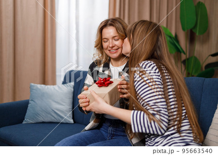 Adult daughter kissing happy mother who sitting on sofa and giving her present 95663540