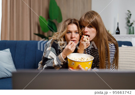 Two women of different ages watch horror movie and eat popcorn while sitting on the couch hugging each other 95663742
