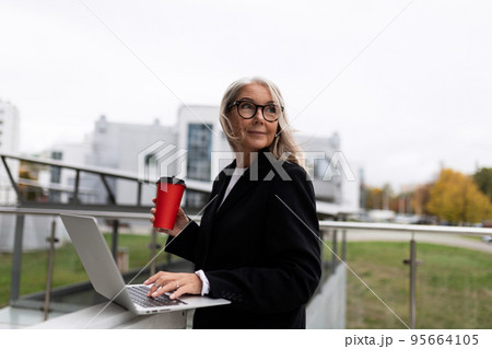Middle aged older business woman works on a laptop and drinks coffee on the office terrace, concept Middle aged older business woman works on a laptop and drinks coffee on the office terrace, concept 95664105