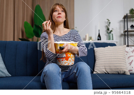 Young caucasian woman spending her leisure at home, watching TV and eating popcorn on sofa. Excited girl with remote control enjoying interesting movie Young caucasian woman spending her leisure at home, watching TV and eating popcorn on sofa. Excited girl with remote control enjoying interesting movie 95664318