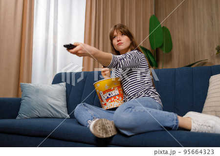 Pretty caucasian woman sitting on sofa, eating popcorn, switching channels, watching tv at home 95664323