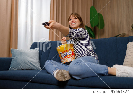 Cheerful young woman with a remote control in hand is watching TV and eating popcorn while spending her free time Cheerful young woman with a remote control in hand is watching TV and eating popcorn while spending her free time 95664324