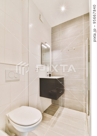Modern flush toilet and ceramic sink installed on white tiled walls near mirror and towel in small restroom at home Modern flush toilet and ceramic sink installed on white tiled walls near mirror and towel in small restroom at home 95667840