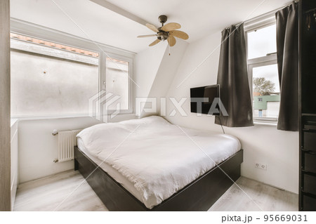 Minimalist interior design of small bedroom with soft bed placed in corner and wooden wardrobe and shelves with lamp illumination 95669031