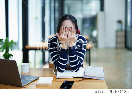 A portrait of a young Asian woman using a computer, wearing headphones and using a notebook to study online shows boredom and pain from video conferencing on a wooden desk in library. 95671589