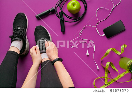 Young woman laces sneakers, preparing for training. Bottle of water, yoga mat, phone, headphones on purple background flat lay top view. 95673579