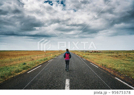 Female tourist running on straight road through farmland and stormy sky in countryside on summer Female tourist running on straight road through farmland and stormy sky in countryside on summer 95675378