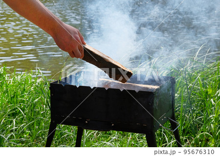 Grill with fuel and charcoal. Mans hand is adding wood into the fire. Barbecue near lake. Grill with fuel and charcoal. Mans hand is adding wood into the fire. Barbecue near lake. 95676310