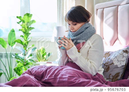 Woman sitting in bed under blanket warming with scarf and hot drink in mug 95676703