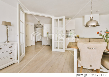 Modern minimalist style interior design of studio apartment with open white kitchen and dining zone with table and chairs illuminated by loft style lamp 95679179