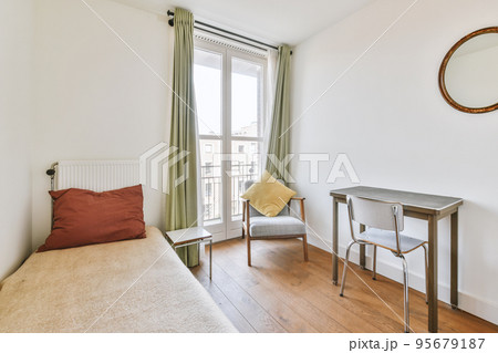 Minimalist interior design of small bedroom with soft bed placed in corner and table with shelves,armchair with lamp illumination Minimalist interior design of small bedroom with soft bed placed in corner and table with shelves,armchair with lamp illumination 95679187