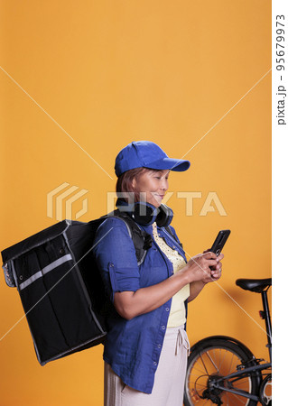 Senior woman deliver carrying take away food backpack checking directions and order details on smartphone. Woman with delivery uniform using bike as transportation. Food service at home Senior woman deliver carrying take away food backpack checking directions and order details on smartphone. Woman with delivery uniform using bike as transportation. Food service at home 95679973