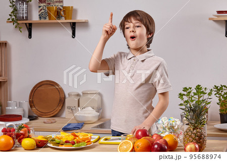 Cute little boy with brown hair is cooking at a kitchen against a white wall with shelves on it. 95688974