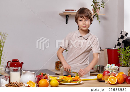 Cute little boy with brown hair is cooking at a kitchen against a white wall with shelves on it. Cute little boy with brown hair is cooking at a kitchen against a white wall with shelves on it. 95688982