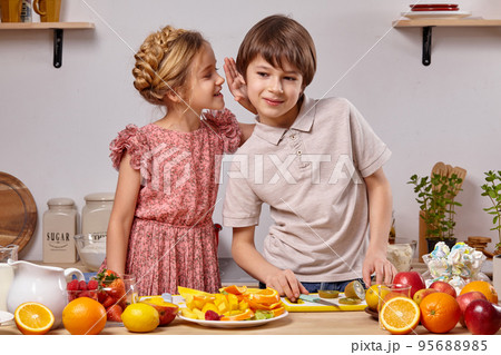 Cute kids are cooking together in a kitchen against a white wall with shelves on it. 95688985