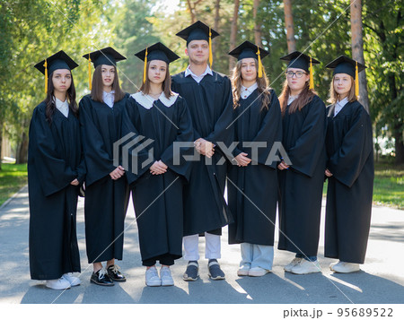Seven graduates in robes stand in a row outdoors. 95689522