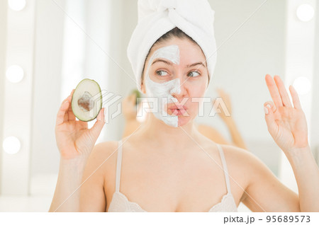A woman with a towel on her hair and in a clay face mask holds an avocado. Taking care of beauty at home 95689573