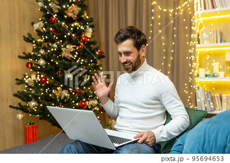 Man at home recording video greetings, Merry Christmas and Happy New Year, sitting on sofa at home near Christmas tree, using laptop for online remote communication with friends, video call. Man at home recording video greetings, Merry Christmas and Happy New Year, sitting on sofa at home near Christmas tree, using laptop for online remote communication with friends, video call. 95694653