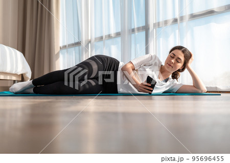 Yoga at home. A young woman in sportswear is lying sideways on a sports Mat and using a smartphone. Windows in the background. Side view. Copy space Yoga at home. A young woman in sportswear is lying sideways on a sports Mat and using a smartphone. Windows in the background. Side view. Copy space 95696455