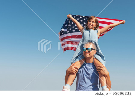 Happy daughter sits on her father's shoulders and holds the American flag above her head, enjoying nature on a sunny day. Free lifestyle. 95696832