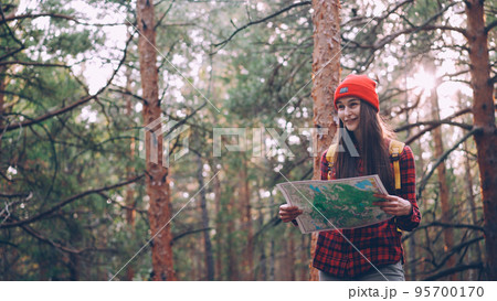 Happy tourist attractive young woman is travelling in forest then looking at map and looking around exploring wild wood. Adventurous people, happiness and sunlight concept. 95700170
