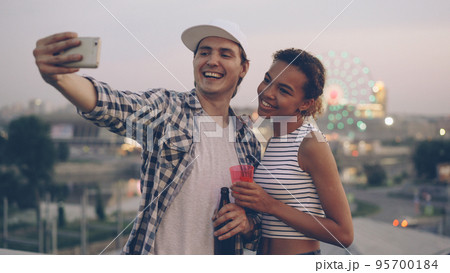 Cheerful multiethnic couple Caucasian young man and African American girl are taking selfie on rooftop using smartphone, laughing and holding drinks with city lights in background. 95700184