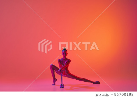 Studio shot of young charming girl, rhythmic gymnast training with sports equipment isolated over pink background in neon light filter 95700198