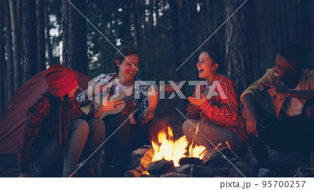 young man tourist is playing the guitar while his friends are singing and laughing sitting around fire in the wood in the evening enjoying nature and good company. 95700257
