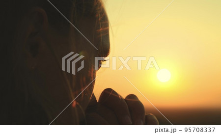 girl prays looking sunset, long hair flying away in glare sunlight rays strong wind, looking at dawn, lonely hike of brave girl, looking into sky with her eyes, believing good. close-up hands face. girl prays looking sunset, long hair flying away in glare sunlight rays strong wind, looking at dawn, lonely hike of brave girl, looking into sky with her eyes, believing good. close-up hands face. 95708337