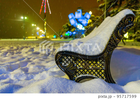 Winter park in the evening covered with snow with a row of lamps View of bench and shining lantern through snowing. Winter night landscape - evening in the night snowy park with lonely benches 95717599