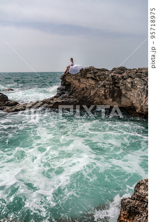 A woman in a storm sits on a stone in the sea. Dressed in a white long dress, waves crash against the rocks and white spray rises. 95718975