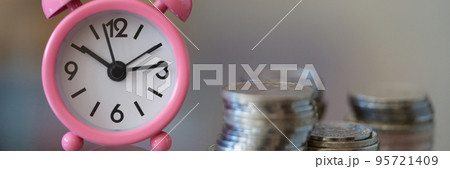 Pink alarm clock standing near pile of coins closeup Pink alarm clock standing near pile of coins closeup 95721409