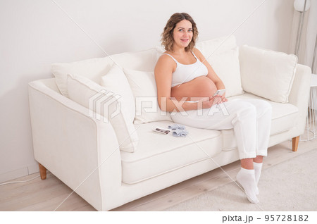 Pregnant woman sits on a white sofa and holds a smartphone. Fetal ultrasonic screening. 95728182