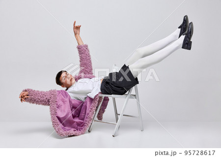 Portrait of stylish boy in pink furry coat, white tights, black shorts lying on chair, posing isolated over grey background. Freedom Portrait of stylish boy in pink furry coat, white tights, black shorts lying on chair, posing isolated over grey background. Freedom 95728617