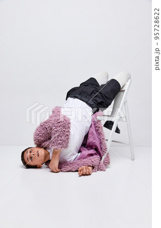 Portrait of stylish boy in pink furry coat, white tights and black shorts lying on chair isolated over grey background. Portrait of stylish boy in pink furry coat, white tights and black shorts lying on chair isolated over grey background. 95728622