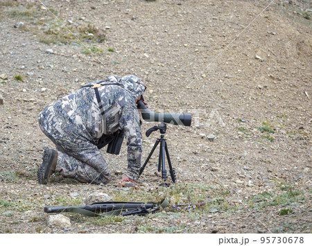 Hunter in camouflage looks carefully through telescope on tripod in mountainous area. 95730678