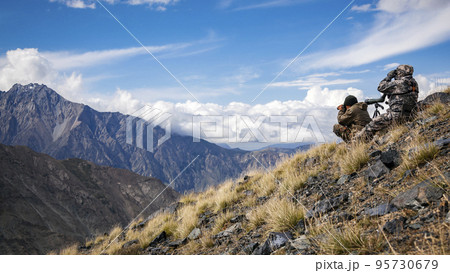 Couple of people dressed in camouflage are carefully examining the mountainous terrain with binoculars. Couple of people dressed in camouflage are carefully examining the mountainous terrain with binoculars. 95730679