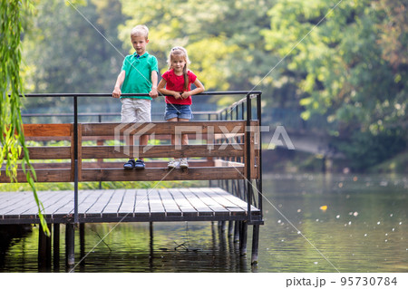 Two children boy and girl standing on wooden deck on a lake shore. 95730784